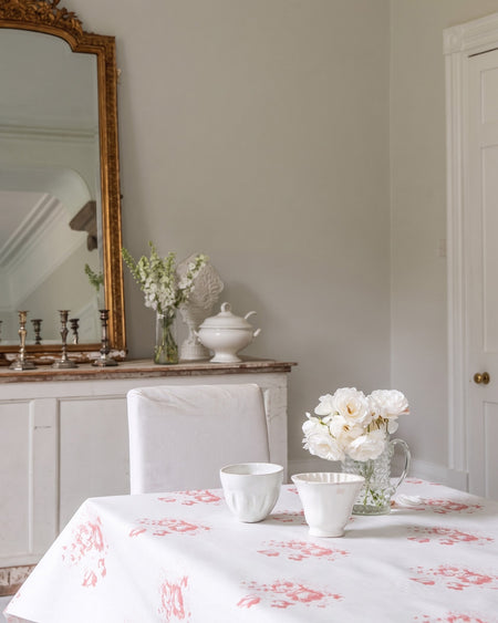 Dining room with floral tablecloth, white chairs, and decorative items on a wooden sideboard.