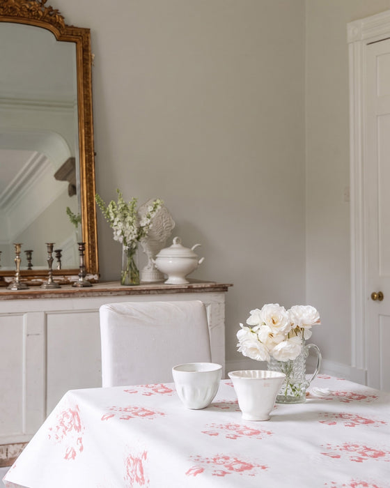 Dining room with floral tablecloth, white chairs, and decorative items on a wooden sideboard.